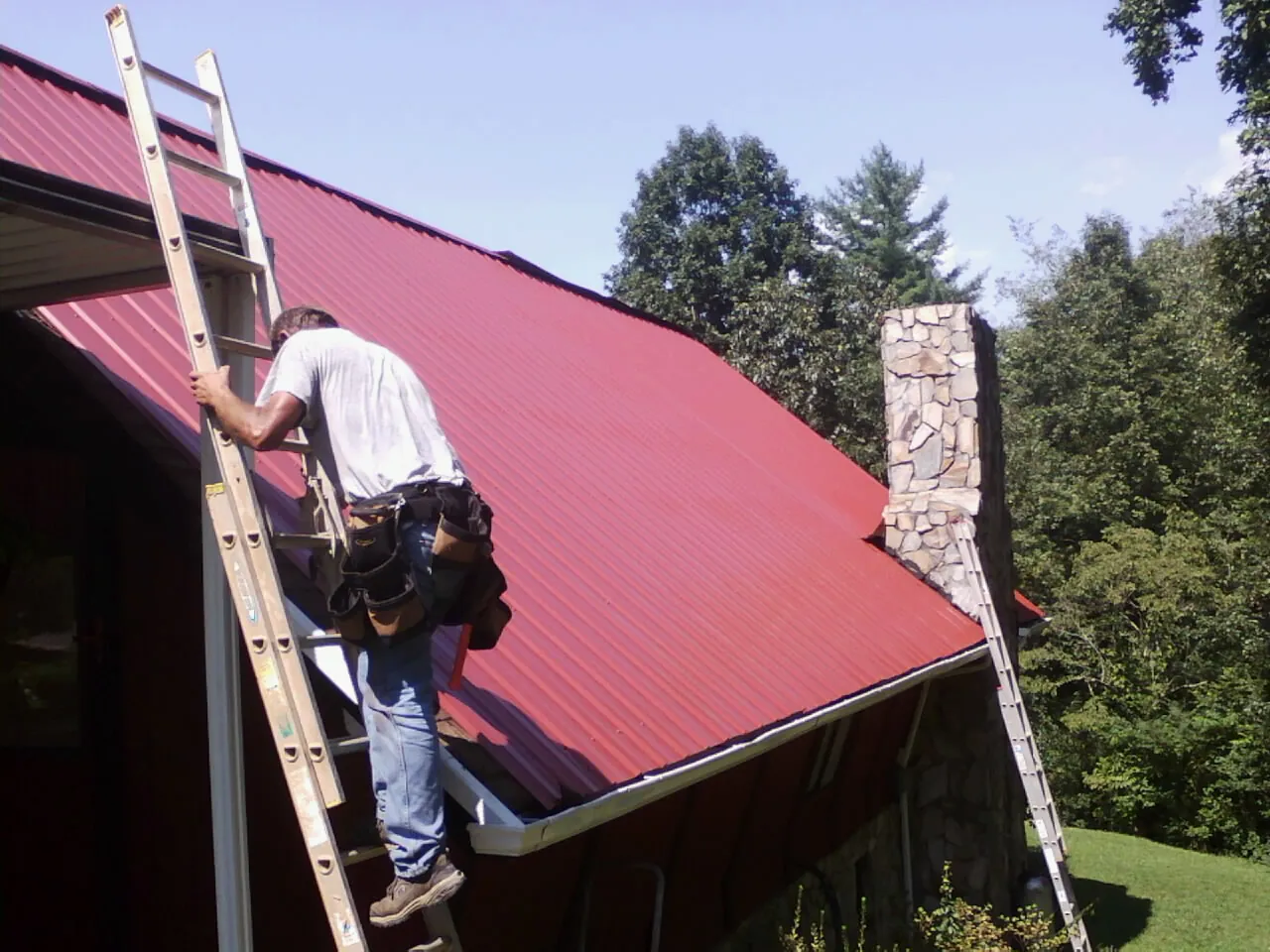 Roofer on a ladder beside a red metal roof and stone chimney — Soffit & Fascia Repair by Roofing Brothers in Iowa City